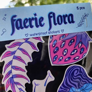 May include: A pack of five waterproof stickers with a faerie flora theme. The stickers feature whimsical designs, including a strawberry with multiple eyes, a mushroom, and a leaf. The stickers are in shades of pink, purple, and blue.