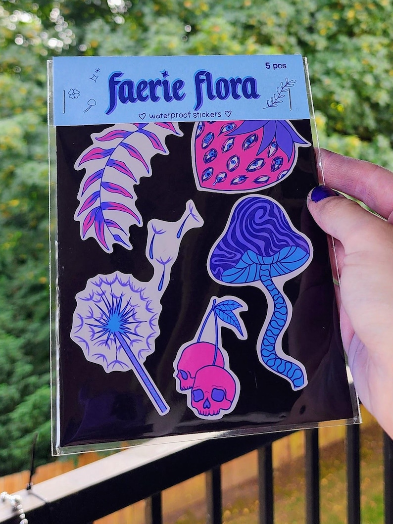 A hand with visible tattoos and blue nail polish holds the sticker pack of colorful plants against a blurred background of green trees and a wooden fence.