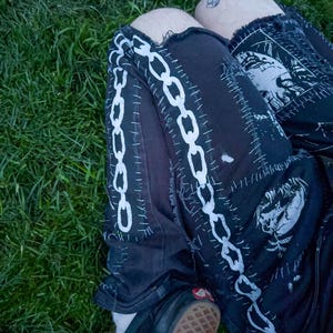 A person wearing black pants with white chain patterns and stitched designs is lying on green grass. A tattoo is visible on their knee.