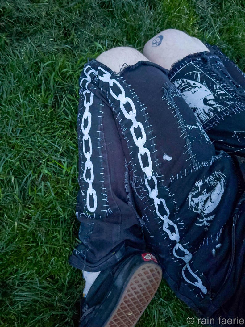 A person wearing black pants with white chain patterns and stitched designs is lying on green grass. A tattoo is visible on their knee.