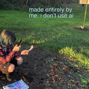 a person with blue hair squats outdoors, taking a photo of a colorful sticker, with a sketchbook on the ground next o them. text reads in the top right reads "made entirely by me, i don't use ai" in light blue. the person has blue hair, visible tattoos, and patch pants. they are outdoors in a field during golden hour.