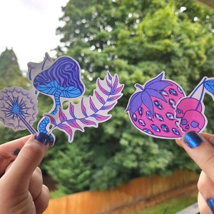 Two hands with metallic blue nail polish hold the stickers against a blurred background of green trees and a wooden fence. The left hand holds three stickers layered together: a purple mushroom, pink-and-purple fern leaf, and a purple dandelion puff with a blue stem. The right hand holds two stickers: a pink strawberry covered in blue eyes with lavender leaves, and two pink skull cherries with a teal leaf.