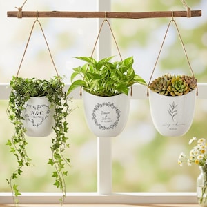 Op de afbeelding: Drie witte hangende plantenbakken met groen, opgehangen aan een houten staaf. Elke plantenbak heeft een uniek zwart ontwerp en tekst. De planten omvatten klimop, pothos en vetplanten.