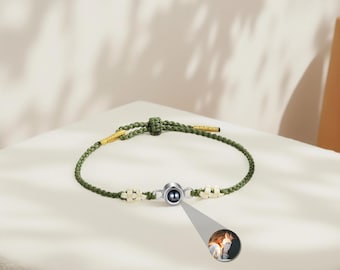 Puede incluir: Un brazalete de cuerda verde con un dije de plata que tiene una perla negra en el centro. El dije tiene acentos de flores blancas a cada lado. El brazalete está atado con un nudo al final.
