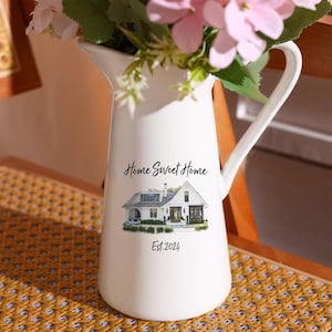 Może przedstawiać: Biały ceramiczny dzbanek z uchwytem, z kompozycją kwiatową. Dzbanek jest ozdobiony ilustracją domu oraz tekstem "Home Sweet Home" i "Est. 2024". Dzbanek stoi na żółtym, plecionym krześle.