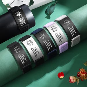 Pode incluir: Seis pulseiras de alerta médica de diferentes cores com uma placa de metal preto gravada com o texto "Devon Black, DOB 09/09/1999, Asma, Equipamento no interior". As pulseiras são feitas de um material macio e elástico e estão disponíveis em preto, azul marinho, roxo, cinza e branco.