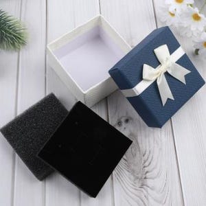 May include: An open white textured gift box and a closed blue gift box with a white ribbon. A black velvet jewelry display card and a black foam insert are next to the boxes. The boxes are on a white wooden surface.