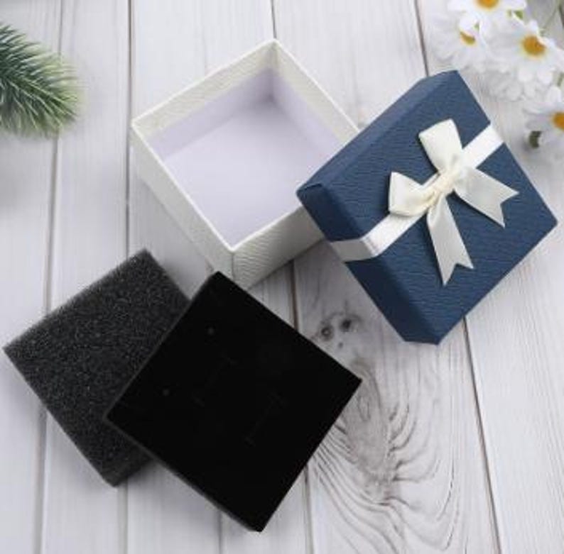 May include: An open white textured gift box and a closed blue gift box with a white ribbon. A black velvet jewelry display card and a black foam insert are next to the boxes. The boxes are on a white wooden surface.