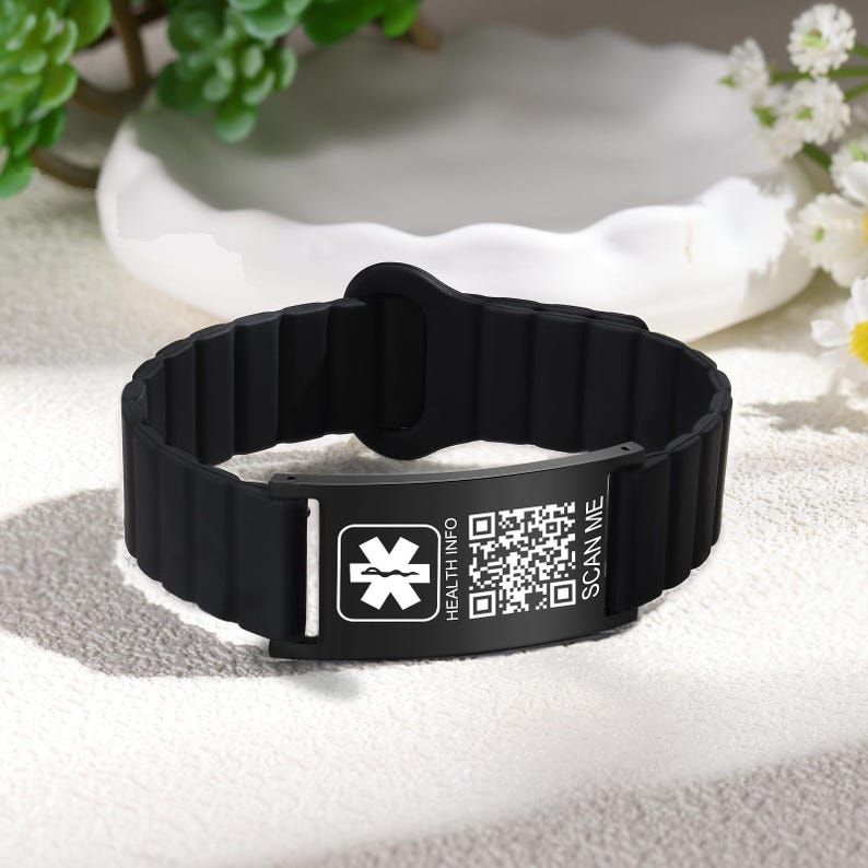 May include: Black medical ID bracelet with a rectangular plate. The plate features a white medical symbol, the words "HEALTH INFO" and "SCAN ME", and a QR code. The bracelet is made of a ribbed, flexible material.