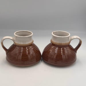 May include: Two ceramic mugs with a wide, rounded base and a narrow neck. The lower portion of each mug features a dark brown glaze with horizontal ribbed textures, while the upper rim and handle are finished in a smooth, light cream glaze.