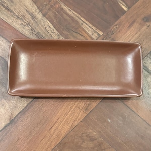 May include: A rectangular ceramic tray with a smooth, solid brown finish. The tray features slightly raised edges and rounded corners, presenting a clean, uniform surface throughout the entire piece, designed for holding small items or serving food.