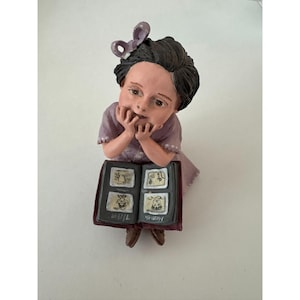 May include: A figurine of a person in a lavender dress, resting their chin on their hands while looking at a photo album. The album is open, displaying several small, framed images. A bow adorns the person's dark hair.