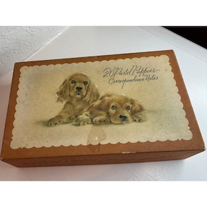 May include: A rectangular brown box featuring a scalloped cream-colored label with an illustration of two golden-brown puppies. The text on the label reads 20 Pastel Puppies Correspondence Notes in a dark brown cursive script.