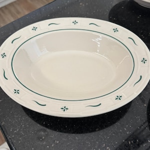 May include: An oval-shaped, off-white serving bowl with a green border and decorative floral and curved line accents. The bowl has a smooth, glossy surface and is likely made of ceramic or porcelain. A classic piece for serving dishes.