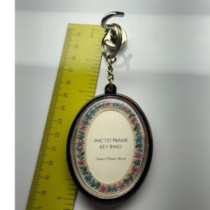May include: An oval photo frame key ring with a black border and a floral pattern around the center insert. The insert features the text 'PHOTO FRAME KEY RING' and '(Insert Photo Here)'. A yellow ruler shows the item is approximately 3 inches long.