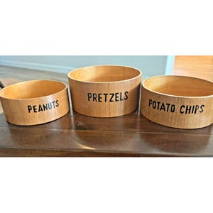 May include: Three wooden serving bowls with black text labels. The bowls are labeled "PEANUTS", "PRETZELS", and "POTATO CHIPS". The bowls are made of light-colored wood and are cylindrical in shape.