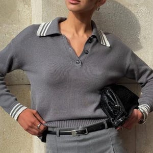 Puede incluir: Un suéter de punto gris con ribetes blancos en el cuello y las mangas. El suéter tiene un cuello abotonado y un ajuste holgado. La modelo lleva un cinturón negro y una falda gris.