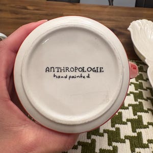 May include: A white ceramic mug with a red rim and handle. The bottom of the mug features the text "ANTHROPOLOGIE hand painted" in black script. The mug is on a green and white patterned surface.