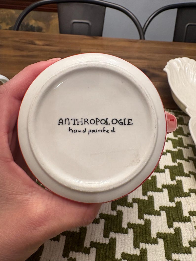 May include: A white ceramic mug with a red rim and handle. The bottom of the mug features the text "ANTHROPOLOGIE hand painted" in black script. The mug is on a green and white patterned surface.