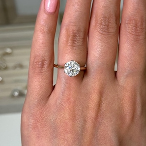 Puede incluir: Un anillo de compromiso de oro con un diamante grande, redondo y transparente. El anillo está engarzado en una fina banda de oro. El anillo está en el dedo de una persona.
