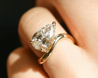 Könnte beinhalten: Ein Verlobungsring mit einem birnenförmigen Diamanten, der in einer goldenen Schiene mit einem einzigartigen, geschwungenen Design sitzt.