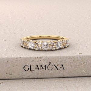 May include: A gold band ring with a series of white, square, and rectangular-cut gemstones. The ring is set on a light-colored surface. The brand name "GLAMOXA" is visible on the surface.