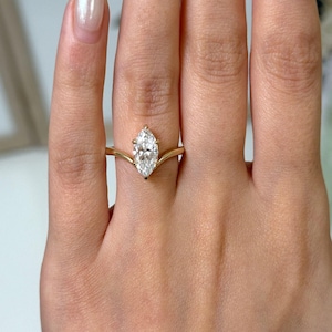May include: A marquise-cut diamond engagement ring with a gold band. The diamond is set in a prong setting, and the band has a unique, curved design. The ring is shown on a person's finger.