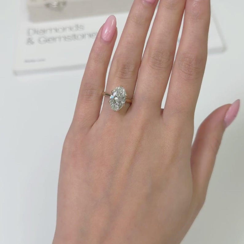 Op de afbeelding: Een gouden verlovingsring met een grote, ovaal geslepen diamant. De ring is gezet op een delicate band en wordt getoond op een hand met verzorgde nagels. De achtergrond bevat een document met de woorden "Diamonds & Gemstone".
