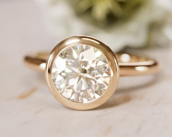 Puede incluir: Un anillo de oro con un diamante grande, redondo y transparente engastado en un bisel. La banda del anillo también es de oro, y el fondo está borroso, con un toque de blanco y verde.