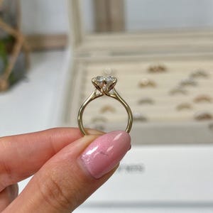 Op de afbeelding: Een gouden verlovingsring met een grote, heldere, rond geslepen diamant. De ring heeft een delicate band en een vier-klauwzetting, die de schittering van de diamant benadrukt. De ring wordt door een hand omhoog gehouden.