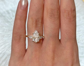 May include: A gold ring featuring a large, pear-shaped diamond center stone, flanked by two smaller diamonds. The ring is set on a delicate gold band, showcasing the brilliance of the diamonds.