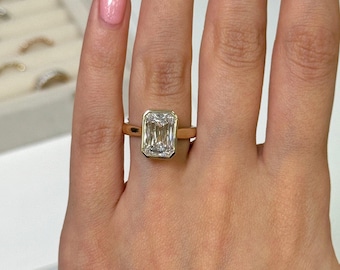 Könnte beinhalten: Ein goldener Ring mit einem großen, rechteckigen, klaren Edelstein. Der Edelstein ist in einer Zargenfassung gefasst, und das Band ist ein schlichtes, poliertes Gold. Der Ring wird an einer Hand gezeigt.
