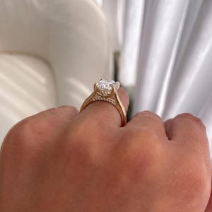 May include: A gold ring featuring a large, clear, cushion-cut center stone held by four prongs. The gallery and the bridge of the ring are accented with a row of small, round, clear stones, adding detail to the polished gold band structure.