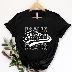 May include: Black t-shirt with the word "Eagles" in a white, retro-style font. The word is stacked with the word "Eagles" repeated above and below. The shirt is hanging on a wooden hanger.