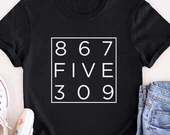 May include: Black t-shirt with a white square graphic that says "867 FIVE 309". The shirt is laid flat on a white surface with a pair of white sneakers and blue jeans.