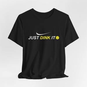May include: Black t-shirt with the text "JUST DINK IT" in white and yellow. The design includes a white swoosh and a yellow pickleball. The shirt is a casual wear item.