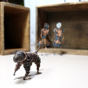 May include: A wire sculpture of a cat walking towards two wire sculptures of people. The sculptures are made of copper wire and have a rustic, aged look.
