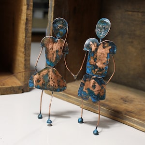 May include: Two copper figures with blue patina stand side-by-side. They are holding hands and have a simple wire frame construction.