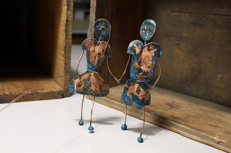 May include: Two copper figures with blue patina stand side-by-side. They are holding hands and have a simple wire frame construction.