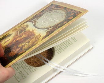 Op de afbeelding: Een close-up van een open boek met een kurken bladwijzer. De boekomslag heeft een gouden achtergrond met een gedetailleerde illustratie van een bos scène en de titel "De oneindige geschiedenis" van Michael Ende.