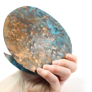 K&ouml;nnte beinhalten: Ein geh&auml;mmerter Kupfer-Wandbehang mit einer blaugr&uuml;nen Patina. Das St&uuml;ck hat eine abgerundete Form und eine strukturierte Oberfl&auml;che.