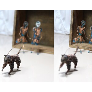 May include: Two wire figures of people with blue and copper paint stand in a wooden box. A wire figure of a cat with copper wire walks in front of the box.