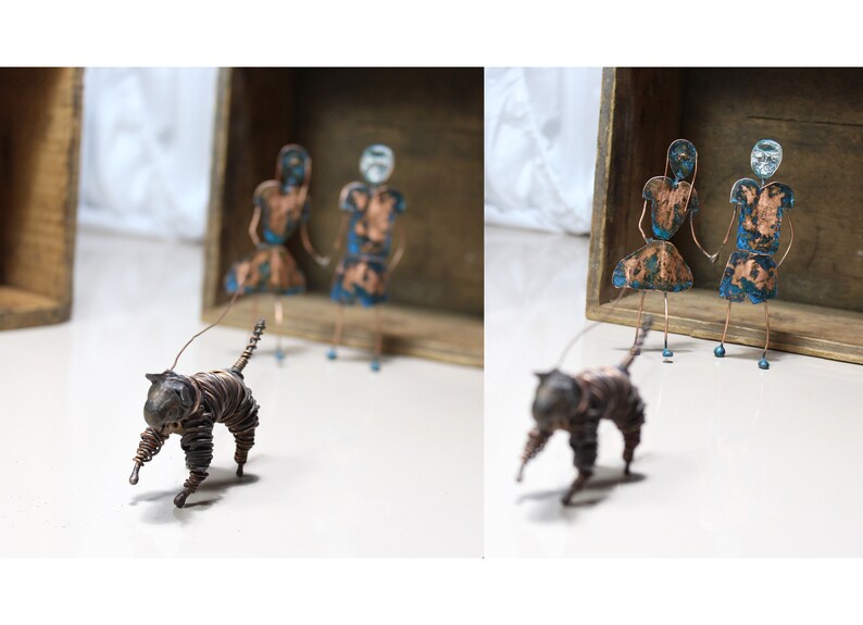 May include: Two wire figures of people with blue and copper paint stand in a wooden box. A wire figure of a cat with copper wire walks in front of the box.