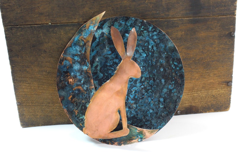 K&ouml;nnte beinhalten: Kupferne Wandkunst mit einer stilisierten Silhouette eines Hasen, der vor einem Halbmond sitzt. Der Mond ist mit einer blauen und gr&uuml;nen Patina texturiert.