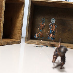 May include: Two small copper figures of people with blue and green accents stand in front of a wooden box. The figures are made of wire and have a rustic, handmade look. A third copper figure, also made of wire, is in the foreground.
