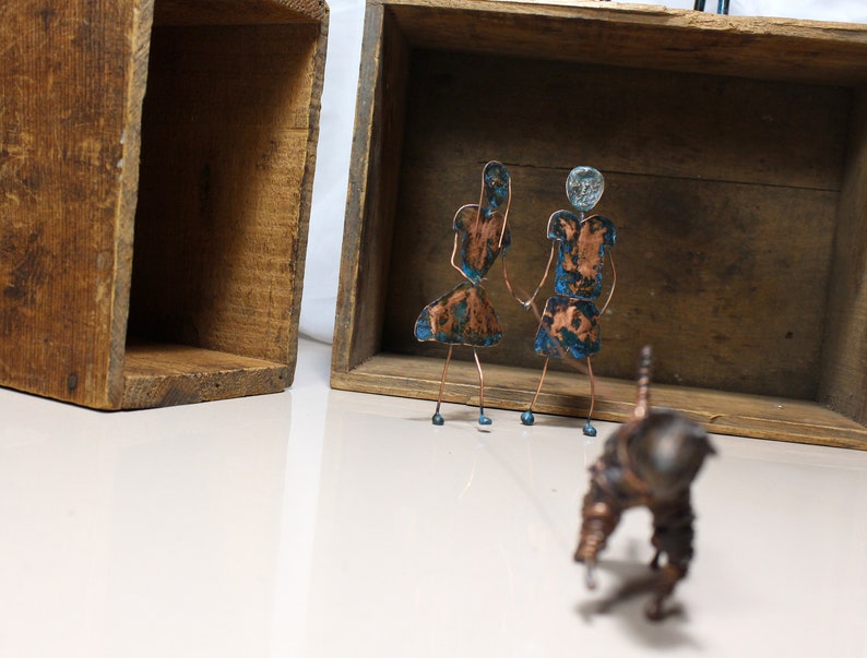May include: Two small copper figures of people with blue and green accents stand in front of a wooden box. The figures are made of wire and have a rustic, handmade look. A third copper figure, also made of wire, is in the foreground.