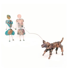 May include: A pair of metal figures, one in blue and one in brown, holding hands and walking a wire-wrapped dog sculpture. The dog is brown and has a metal tag that says "Sam".