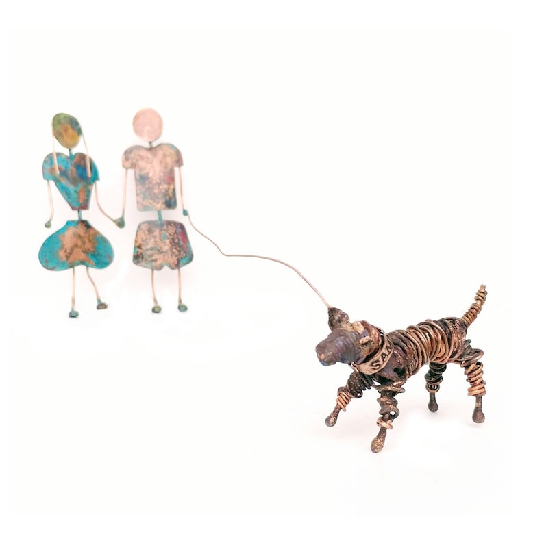 May include: A pair of metal figures, one in blue and one in brown, holding hands and walking a wire-wrapped dog sculpture. The dog is brown and has a metal tag that says "Sam".