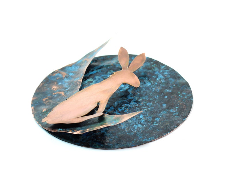 K&ouml;nnte beinhalten: Eine Kupferskulptur eines Kaninchens auf einer blauen und kupferfarbenen, strukturierten Scheibe.