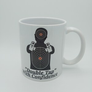 K&ouml;nnte beinhalten: Eine wei&szlig;e Keramiktasse mit einer schwarzen Silhouette einer Person, die zwei Pistolen vor einer Zielscheibe h&auml;lt. Der Text "Double Tap" und "With Confidence" ist unter der Silhouette aufgedruckt.